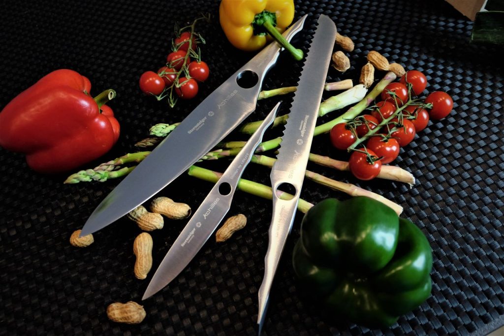 A collection of chefs knives with peppers, tomatoes, and peanuts on a black surface.