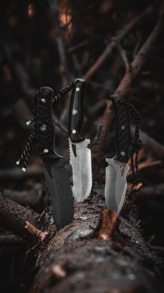 Three survival knives stuck into a tree trunk in a dark, moody forest setting.